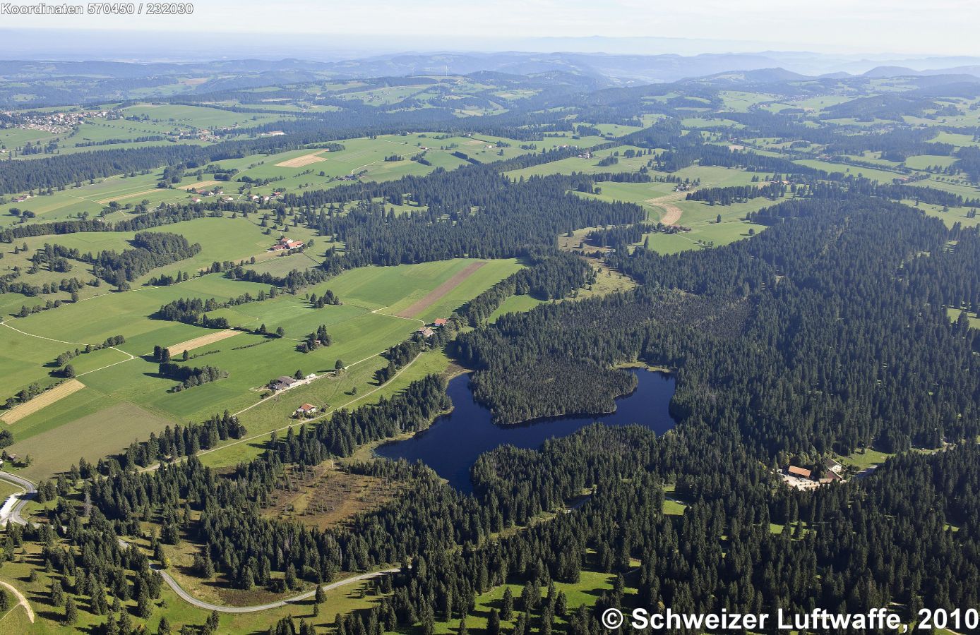 Freiberge (Jura), Blickrichtung NE. Im Zentrum liegt der Etang de la Gruère. Kleiner Weiler unten rechts im Wald: Moulin de la Gruère. Siedlung links oben: Montfaucon (993 m.ü.M.) Dahinter in der Schlucht: Doubs.